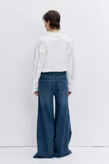 Camisa de algodón color blanco con detalles de encaje broderie y alforzas verticales. Presenta cuello alto con volados, cierre frontal de botones y mangas largas con puños acampanados.