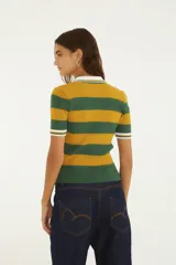 Remera polo de punto acanalado con diseño de franjas horizontales en verde y amarillo. Presenta cuello tipo polo con ribete blanco, mangas cortas con puños rayados, cierre frontal parcial con botones y parche circular bordado de la marca en el pecho.
