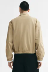 Campera tipo bomber de sarga de algodón en color beige, con cuello inglés, cierre frontal metálico y bolsillos diagonales ribeteados. Presenta terminaciones con elástico en los puños y el ruedo.