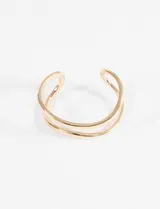 Pulsera tipo cuff rígida de metal dorado, con diseño abierto y dos bandas curvas entrelazadas de grosor uniforme.