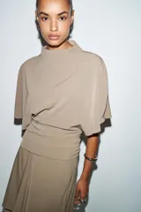 Top fluido color beige, con cuello perkins y mangas por debajo del codo. Bajo redondeado.