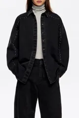 Camisa de corte oversize confeccionada en mezcla de algodón, con cuello camisero, cierre frontal de botones y manga larga. Presenta detalles de remaches metálicos a lo largo de las costuras de las mangas y el cuello.