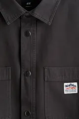 Sobrecamisa negra de lona gruesa de algodón, con cuello, tapeta con botones a presión y canesú en la espalda. Tiene mangas largas con botón a presión en los puños y dos bolsillos superiores de parche. El bajo es levemente redondeado y el corte es oversized.