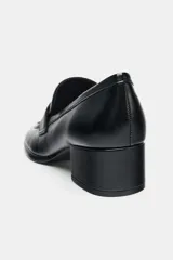 Mocasín de mujer en color negro con acabado liso, diseño de punta cuadrada y tacón en bloque. Presenta un detalle metálico decorativo en el antifaz frontal.