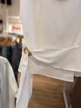 Blusa blanca de diseño cruzado sin mangas, con escote bote y un lazo lateral con hebilla para ajustar en la cintura.