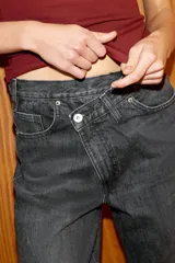 Jeans de tiro alto, color gris oscuro lavado, con pierna ancha extra larga (full length) y diseño de cintura cruzada asimétrica con cierre frontal de cremallera y botones metálicos.