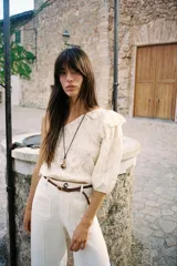 Top corto asimétrico color blanco roto, con un solo hombro descubierto y manga abullonada corta en el otro lado. Presenta bordados perforados (ojales) y un volante en el hombro descubierto. El bajo y el puño de la manga terminan en elástico.