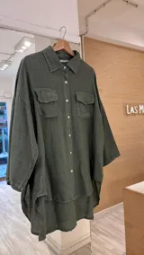 Camisa verde militar de lino, de corte amplio, con cuello clásico, cierre frontal con botones de nácar, dos bolsillos delanteros con solapa, mangas largas con puños abotonados, bajo asimétrico con aberturas laterales y espalda más larga que el frente.