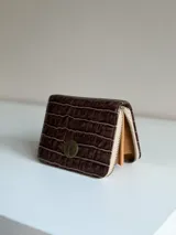 Billetera pequeña marrón con textura de cocodrilo, cierre de cremallera perimetral y detalle de chapa metálica redonda en la solapa frontal.