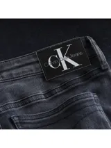 Jeans de mezclilla gris oscuro lavado, corte skinny y talle medio, con una banda lateral vertical que lleva el texto "Calvin Klein Jeans" repetido.