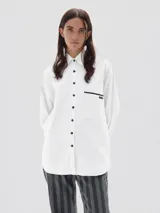 Camisa de corte oversized confeccionada en tela oxford de algodón. Presenta cuello clásico, cierre frontal con botones negros a contraste y un bolsillo en el pecho con detalle de ribete negro y etiqueta de marca.