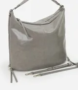 Bolso grande tipo hobo desestructurado, color gris claro con acabado ligeramente brillante o envejecido. Cuenta con dos asas cortas y una correa larga desmontable. Presenta detalles de cierres o tiradores decorativos en los laterales y cierre principal superior con cremallera.