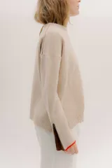 Buzo de lambswool color beige, con escote redondo y detalle en V. Presenta cierres metálicos en los puños que revelan un interior en contraste de color marrón y ribete rojo. Tiene tajos laterales para mayor comodidad y un fit amplio.