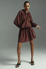 Vestido túnica corto color marrón en mezcla de lyocell y viscosa con leve brillo. Modelo oversized con cuello redondo con ribete trenzado grueso, escote en V con tiras finas, mangas globo largas con abertura en los hombros, elástico en los puños y cintura fruncida con vuelo en la falda.
