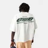 Remera oversized color blanco crema, con cuello redondo y mangas cortas. Presenta un estampado en la espalda con el logo de "Eme Studios" en verde y texto "Building a community through gratitude".