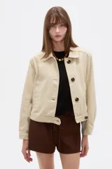 Chaqueta corta de algodón color beige, con cuello camisero, bolsillos laterales y cierre frontal con botones oscuros.