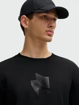 Camiseta negra de corte relajado con cuello redondo y manga corta. Presenta un diseño tridimensional en el pecho que simula un pliegue o estructura geométrica en tono negro brillante o texturizado.