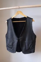 Chaleco reversible confeccionado en cuero genuino. Presenta un lado de piel de oveja gris con bolsillos de cuero negro y un lado de cuero liso gris. Diseño sin mangas con cierre frontal.