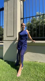 Vestido largo color marrón, con escote recto y tirantes finos. Presenta un diseño con nudo frontal que crea un efecto drapeado y una abertura central que deja ver una capa inferior más corta.