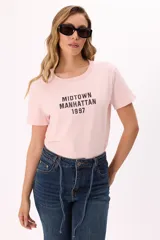 Remera de algodón color rosado con cuello redondo y mangas cortas. Presenta un estampado frontal en color negro con el texto 'MIDTOWN MANHATTAN 1997' en tipografía estilo varsity.