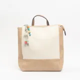Mochila de diseño bicolor en tonos crema y beige, con cierre superior, asa de mano y detalle de colgante con cuentas coloridas.