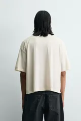 Camiseta de corte holgado y estilo cropped, color beige con efecto lavado. Presenta cuello redondo, mangas cortas y una estampa frontal con texto en azul que simula trazos de rotulador.