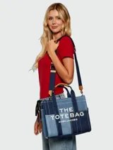 Cartera tipo tote bag Marc Jacobs modelo The Small Traveler Tote, confeccionada en denim con diseño de paneles en distintos tonos de azul. Cuenta con asas superiores, correa de hombro ajustable y extraíble, y el texto "THE TOTE BAG MARC JACOBS" estampado en la parte frontal.