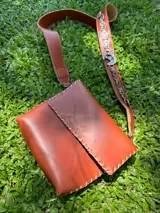 Cartera de cuero de formato rectangular con solapa frontal y costuras vistas artesanales en el contorno. Cuenta con una correa larga de cuero que incorpora una aplicación de cadena metálica.