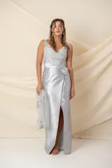 Vestido largo de fiesta en color gris plata, con cuerpo de tul bordado con motivos florales y escote en V. Presenta una cintura drapeada con detalle de moño lateral y falda cruzada con abertura frontal. Incluye un chal de tul a juego con detalles bordados en los extremos.