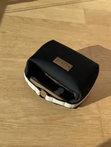 Necessaire de neopreno color negro con cierre superior, forro interno y compartimento. Presenta una manija lateral de cinta color beige con detalles en negro y un aplique de cuero sintético con el logo de la marca.