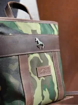 Mochila mini Sierra Mora de diseño estructurado, confeccionada en lona con estampado camuflado y detalles en cuero marrón. Cuenta con tiras de cuero verticales, asa superior, dije metálico en forma de caballo y un parche con el logo grabado de la marca.