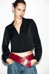 Camisa corta de tejido satinado color marrón, con cuello solapa, escote en pico y manga larga. Presenta cierre frontal con botones forrados a tono.