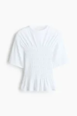 Remera de algodón blanca con hombros levemente caídos, cuello redondo y un amplio sector central con fruncido elástico que finaliza en un pequeño volante en el ruedo.