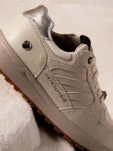 Sneaker urbana de edición limitada inspirada en Star Wars, color off-white con detalles en plateado. Presenta el texto 'JOIN THE RESISTANCE' en el lateral, parche de Star Wars en la lengüeta y dije metálico en los cordones. Suela de plataforma en tono arena con base de goma dentada.