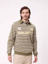 Polo de manga larga con diseño a rayas horizontales en color verde oliva y blanco. Presenta cuello tipo polo con cierre de botones, estampado frontal con el número 85, logo de la marca Rusty y escudo tipo universitario.