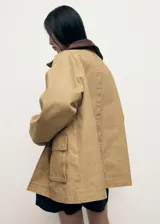 Anorak de estilo oversize en color tostado, con cuello camisero de pana en contraste. Presenta cierre frontal mediante ganchos metálicos y dos bolsillos de parche con solapa en la parte delantera.
