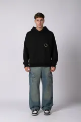 Buzo canguro oversize de frisa de algodón premium en color azul grisáceo. Presenta un diseño con recortes frontales, bolsillo tipo canguro y un pequeño logo circular estampado en el pecho. Cuenta con puños y ruedo acanalados.