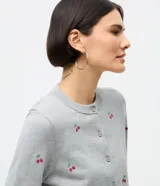 Cardigan corto de punto color gris, con cuello redondo, mangas largas y cierre frontal con botones. Presenta un diseño ajustado al cuerpo con bordados de cerezas distribuidos por toda la prenda.