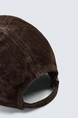 Gorra de béisbol color marrón oscuro, confeccionada en material con textura de ante o nobuk. Presenta visera curva y ojales metálicos a los lados para ventilación.