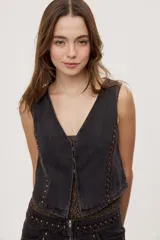 Chaleco corto de denim negro con escote en V y diseño sin mangas. Presenta terminación lavada y detalles laterales con entramado y tachas metálicas que aportan un toque distintivo.