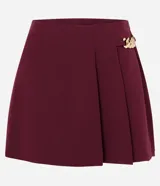 Short pollera de sastrería marca Cortelle, color bordó, confeccionado en crepé. Presenta sobreposición frontal efecto pollera con pliegues y detalle de cadena metálica decorativa en el lateral.