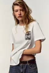 Camiseta blanca de manga corta y cuello redondo, con aplicación de organza y texto negro a contraste en el pecho.