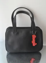 Cartera negra de cuero sintético con doble asa corta y dije colgante con la palabra 'Pineapple'.