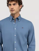 Camisa de hombre color azul, con cuello buttondown, cierre frontal con botones y logo bordado en el pecho. Presenta un acabado vintage logrado mediante el proceso de teñido en prenda.