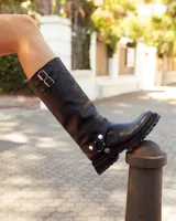 Bota de caña alta confeccionada en cuero negro, con suela track de goma. Presenta un diseño versátil con dos hebillas ajustables en la parte superior de la caña y una correa decorativa con tachas metálicas y argollas en la zona del tobillo.