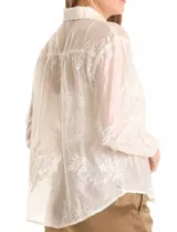 Camisa de tencel color blanco roto, semitransparente, con bordados florales en blanco. Tiene cuello con solapa, cierre frontal con botones decorados y un bolsillo en el pecho.