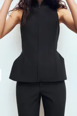 Chaleco entallado de color negro con cuello alto y cierre frontal con cremallera oculta. Presenta un diseño sin mangas con costuras verticales que definen la silueta y un bajo con ligero volumen acampanado.