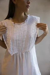 Vestido mini blanco de silueta amplia y recta, con escote redondo, volados en los hombros con detalles de puntillas, pechera con tablas y guarda bordada, y terminación desflecada en el ruedo.