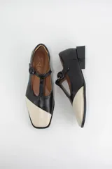 Zapatos tipo Mary Jane bicolor en negro y crema, confeccionados en cuero. Presentan una tira en forma de T con cierre de hebilla lateral y un taco cuadrado bajo de 3 cm.