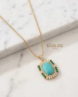 Collar de cadena fina con baño de oro y dije ovalado con piedra central color turquesa, rodeada por un marco de circonias blancas y pequeñas piedras verdes.